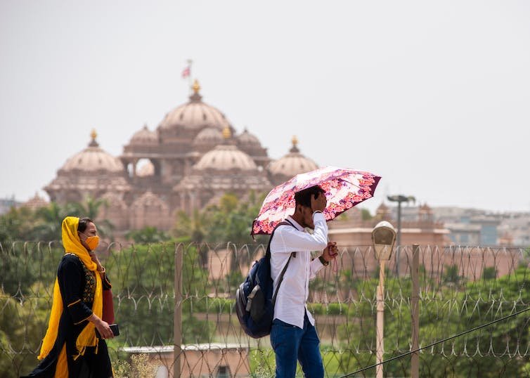 दो लोग, एक छाता लिए हुए, एक गर्म दिन पर पृष्ठभूमि में एक हिंदू मंदिर के साथ चल रहा है।