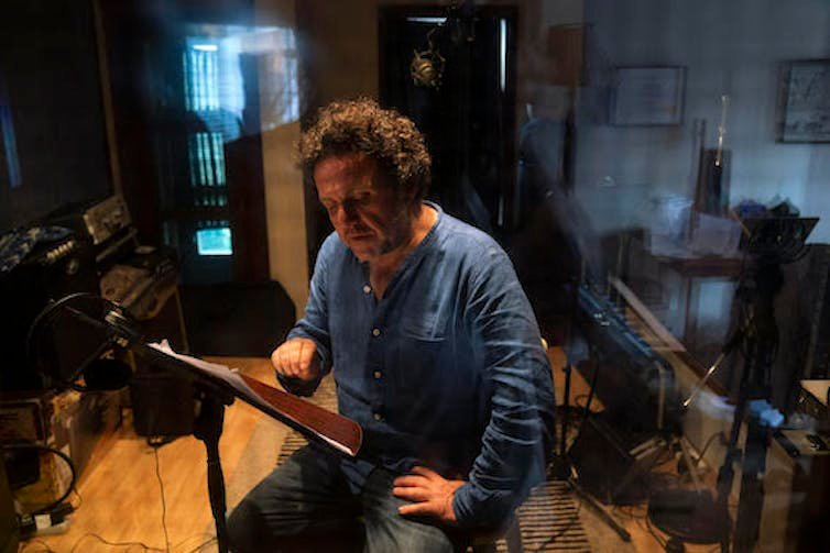 एक आदमी एक लकड़ी के फ़र्श वाले स्टूडियो में एक संगीत स्टैंड, संगीत उपकरण पर कागजात से पढ़ने वाली कुर्सी पर बैठता है।